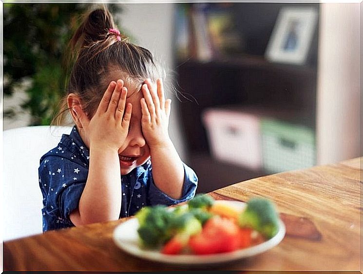 little girl holding her eyes with the plate in front of her with food